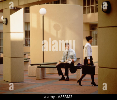 Man with newspaper notices passing woman while seated on bench in mall. Stock Photo
