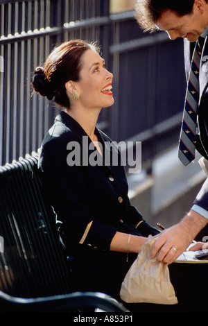 Close-up of smartly dressed woman sitting on bench smiling up at man in suit standing outdoors in urban setting. Stock Photo