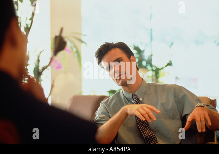 Close-up of seated man in shirt and tie. Causal indoor setting with back of man's head and shoulder out of focus. Stock Photo