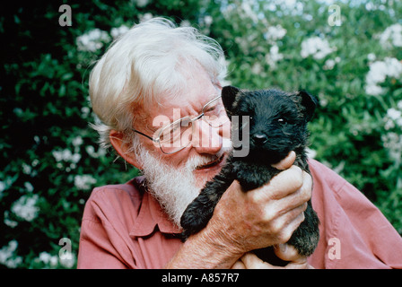 Outdoor garden portrait of an old man with grey hair and beard holding a black puppy dog. Stock Photo