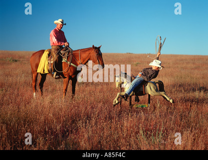 Young cowboy rides his electric horse while an older cowboy and his real horse look on Stock Photo