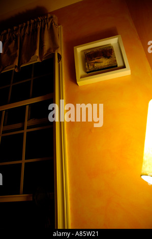 Orange bedroom wall glowing in the light from an incandescent bulb with white window frame and lattice work to left of artwork Stock Photo