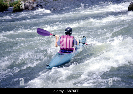 A man in a light blue kayak paddling down a churning river while wearing a life vest and helmet Stock Photo