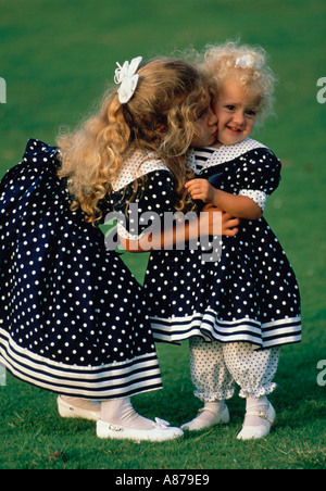 A blond girl hugs her younger sister and gives her a kiss on the cheek The girls are wearing blue and white polka dot dresses Stock Photo