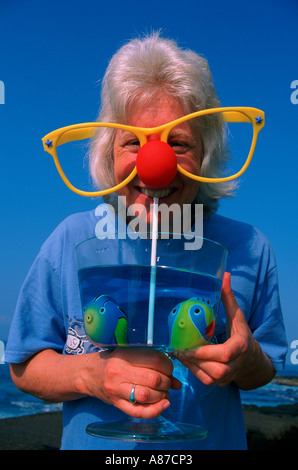 A middle aged woman wearing oversized clown glasses and sipping from a glass with fake plastics fish taken in Kona Hawaii Stock Photo