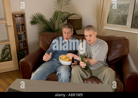 Model Released. Two football fans sitting on sofa watching TV, shouting and jeering whilst eating crisps and drinking beer. Stock Photo