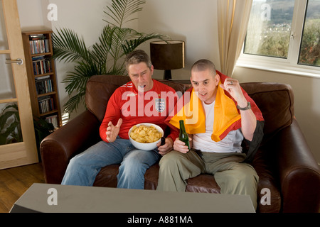 England and German football fans sitting on sofa watching TV. Shouting and jeering whilst eating crisps and drinking beer. Stock Photo