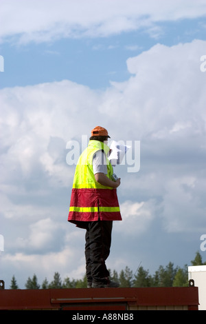 Foreman standing against cloudy sky at the top of a container wearing colorful safety vest and checking the construction plans , Finland Stock Photo