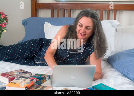 View of youthful mature woman with long grey hair working on her laptop while relaxing in bed Stock Photo