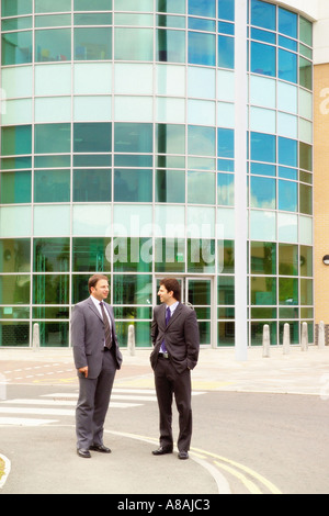 Two Caucasian businessmen in suits standing outside a modern office building with a curved glazed facade Stock Photo