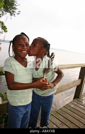 Two Afro American young girls hugging and kissing on the dock near lake. Stock Photo