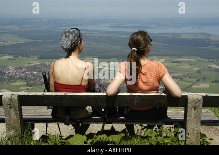 Back of two young women sitting on a bench with a view onto Chiemsee at Hochgern mountain Chiemgau Bavaria Stock Photo