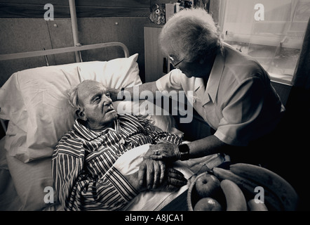 CORONAVIRUS Visiting carer wife companion comforts elderly gentleman in his care home bed Black and White toned treatment Stock Photo