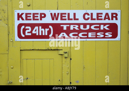 Red and white sign with bold letters stating Keep well clear 24 hour Truck access on a yellow wooden door Stock Photo