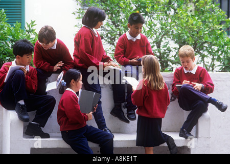 Group of junior multiracial school children sitting studying interacting talking together outside on a white wall in the playground Stock Photo