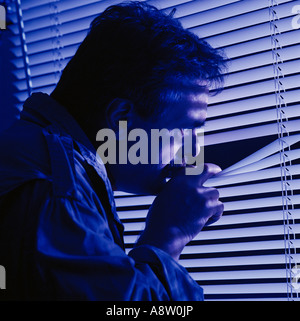 Indoor close-up of frightened man looking out of window through venetian blinds. Stock Photo