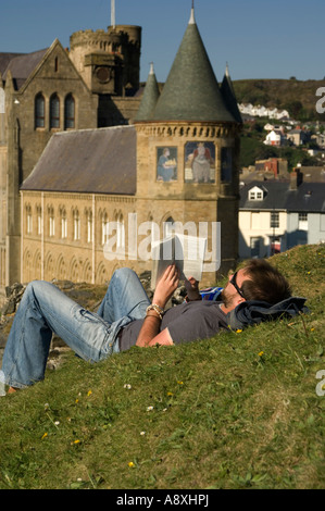 young man student lying on grass reading a book with the University of Wales Aberystwyth Old College in background, summer UK Stock Photo