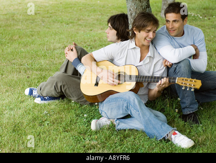 Young male friends sitting near tree, playing guitar Stock Photo