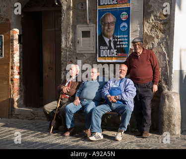 Four old Italian men on bench under political poster outside Tobacco shop Cefalu Sicily Italy Stock Photo