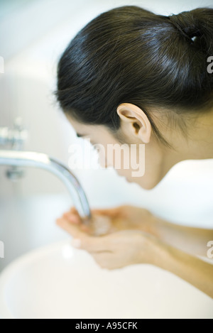 Young woman leaning over sink, hands cupped under faucet Stock Photo