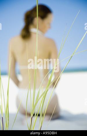 Woman in bikini kneeling on beach, focus on sea oats in foreground