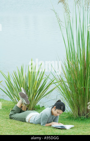 Young woman lying on grass near body of water, reading
