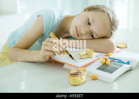 Girl sitting at table doing homework, resting head on arm, doodling, food scattered on books Stock Photo