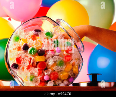 A child hand reaches into a colorful candy jar with colorful balloons in the background Stock Photo