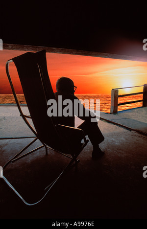 An elderly man sits in a beach chair looking out from under the boardwalk at the sunset on the ocean Stock Photo