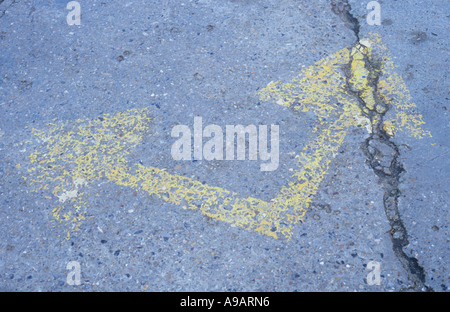 Two well worn yellow arrows at right angles to each other painted on a cracked tarmac road surface Stock Photo