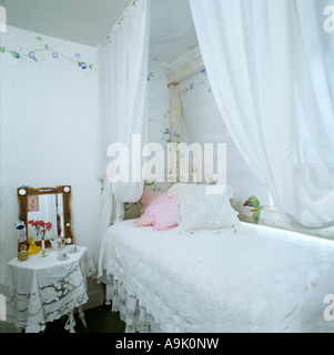 White voile drapes above bed with white lace cover in small white bedroom with stencilled blue floral border on the walls Stock Photo