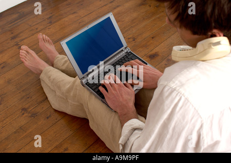 man using a laptop with a phone tucked under his ear Stock Photo