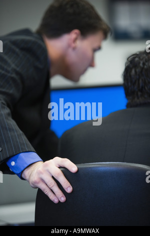 Business man with hand resting on back of chair leaning over computer monitor with colleague Stock Photo