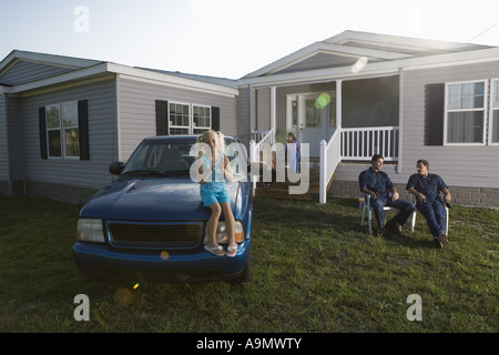 Girl sitting on hood of old truck playing patty-cake, family in background Stock Photo