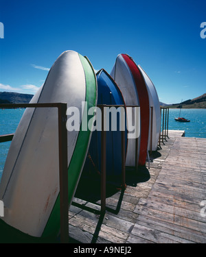 Dinghies standing up on dock at Okains Bay with a sailboat in distance in harbour community of Sumner near Christchurch Stock Photo