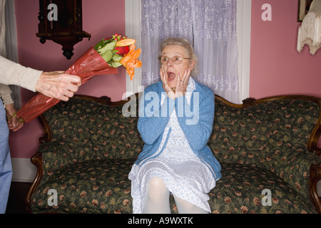 Surprised and happy senior woman sitting on the sofa handed bouquet of flowers Stock Photo