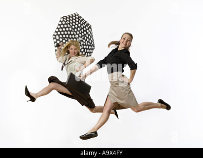 two young women leaping in the air wearing office clothes one with a brief case, the other with an umbrella. Fantasy commute. Stock Photo