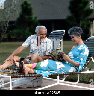 Older couple brightly dressed sitting in deck chairs having an animated conversation while she looks over a magazine Stock Photo