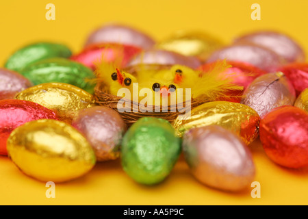 Foil covered Easter egg chocolate confectionary with bright yellow novelty chick toys in wicker basket on a yellow background Stock Photo