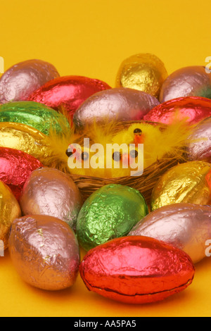 Foil covered Easter egg chocolate confectionary with bright yellow novelty chick toys in wicker basket on a yellow background Stock Photo