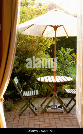 View through French windows onto a summer courtyard garden, featuring a small round table and chairs under a cream-coloured parasol. Stock Photo