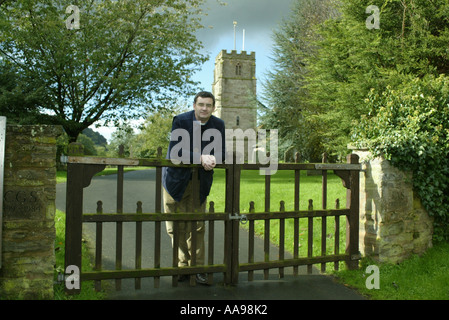 A rural vicar leans on the gate to the country church in Herefordshire, England. Stock Photo