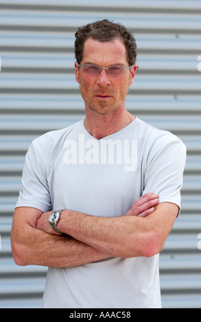middle aged caucasian man wearing a light grey T-shirt and glasses stands with his arms folded in front of a metal wall Stock Photo