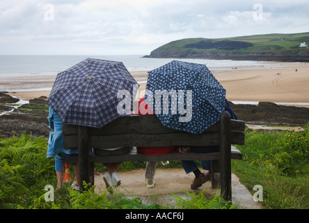 Two people sat on wooden bench each holding a rain brolly over them looking at view of Croyde Bay Devon England Stock Photo