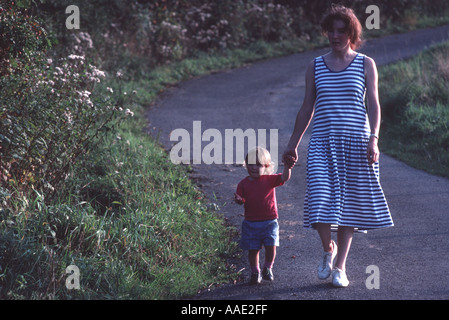 Woman holding hands with her small daughter aged about two years walking towards camera on a winding woodland path in backlight. Stock Photo