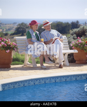 mature couple relaxing reading a book by the pool, the  sea in background Stock Photo