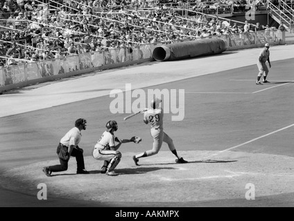 1970s SIDE VIEW OF BATTER COMPLETING SWING WITH CATCHER & UMPIRE BEHIND HIM AT PROFESSIONAL BASEBALL GAME Stock Photo