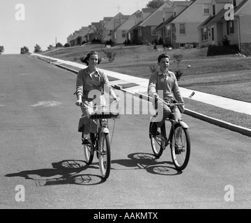 1950s TEEN BOY GIRL COUPLE RIDING BIKES DOWN RESIDENTIAL STREET Stock Photo