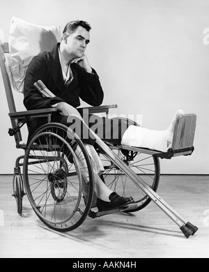 1940s MAN IN WHEELCHAIR WITH CAST ON LEFT LEG HOLDING CRUTCHES Stock Photo