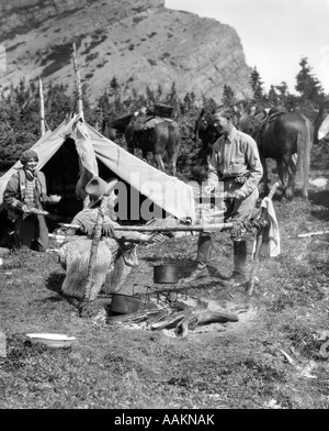 1920s 1930s TWO MEN AND ONE WOMAN EATING A MEAL AROUND A CAMPFIRE WITH A TENT AND HORSES AT BAKER LAKE ALBERTA CANADA Stock Photo